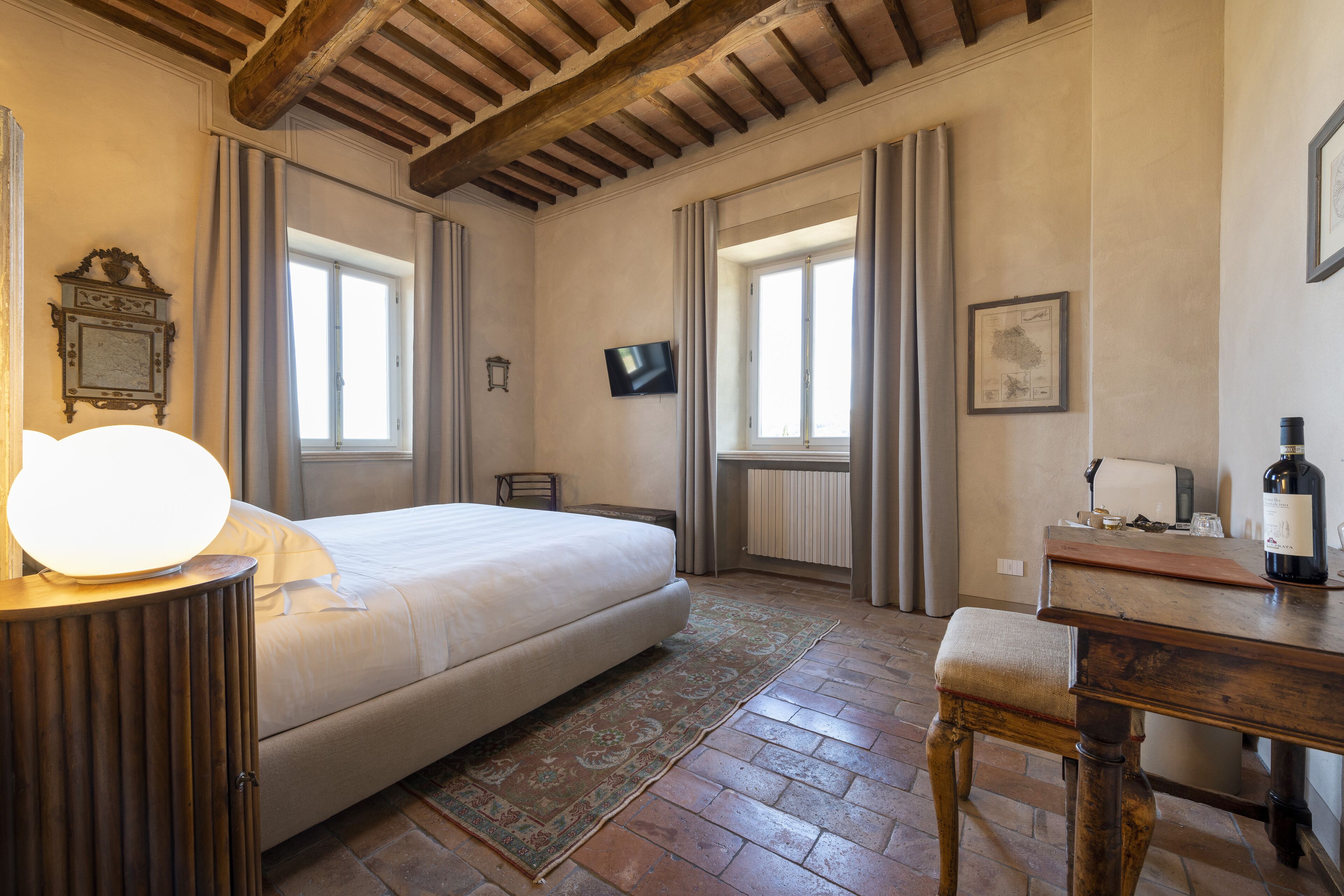 Un dormitorio con cama, una mesita de noche con lámpara, un banco de madera, un televisor en la pared y una ventana con cortinas.
