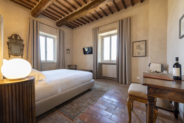 A bedroom with a bed, a bedside table with a lamp, a wooden bench, a TV on the wall, and a window with curtains. - Villa Le Prata - Residenza del Vescovo (Montalcino)