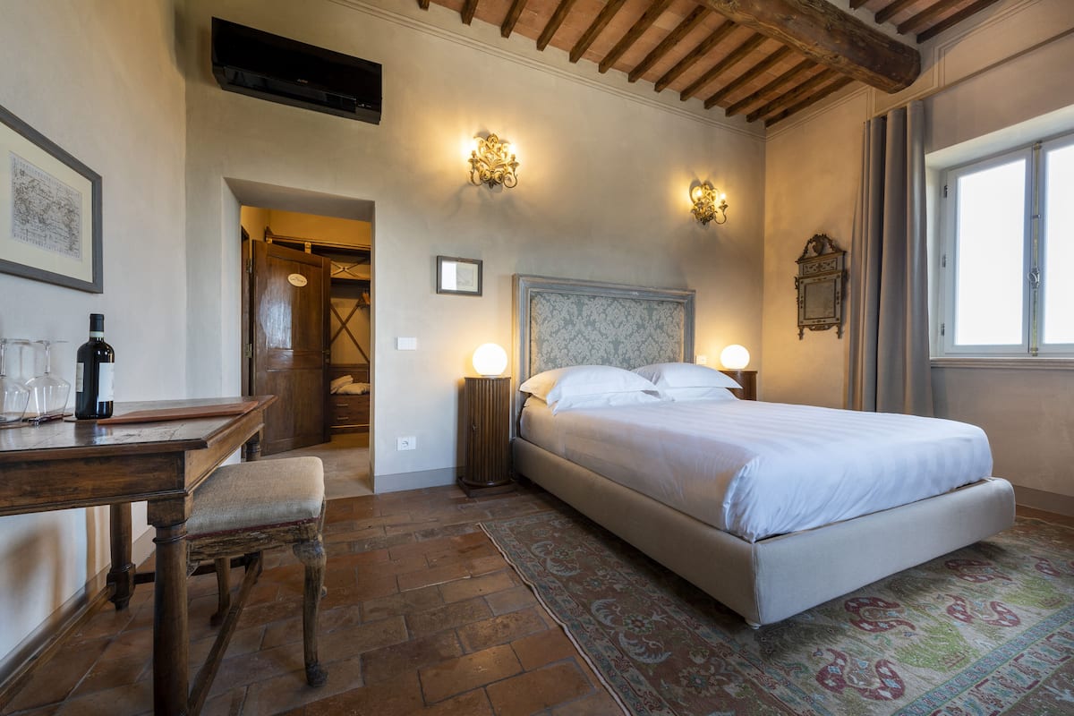 A bedroom with a bed, bedside tables, a desk, and a chair. There is a window and a door leading to another room.