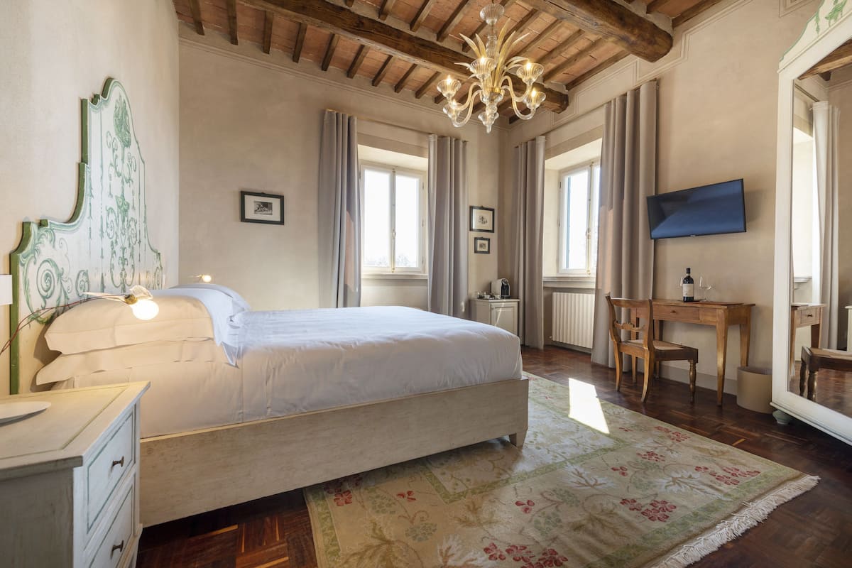 A bedroom with a bed, a desk, a chair, a chandelier, and a large mirror.