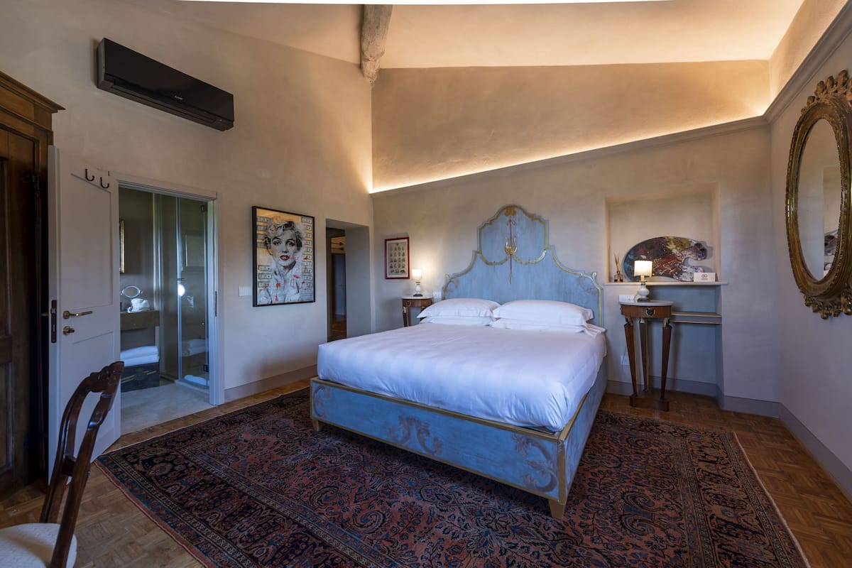 A bedroom with a large bed, a wooden chair, a mirror, and a small table with a lamp.