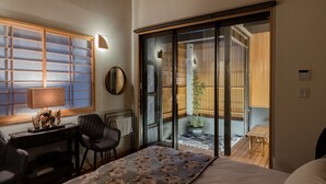 A bedroom with a bed, a desk, a chair, a mirror, and a sliding glass door leading to a balcony with a potted plant.