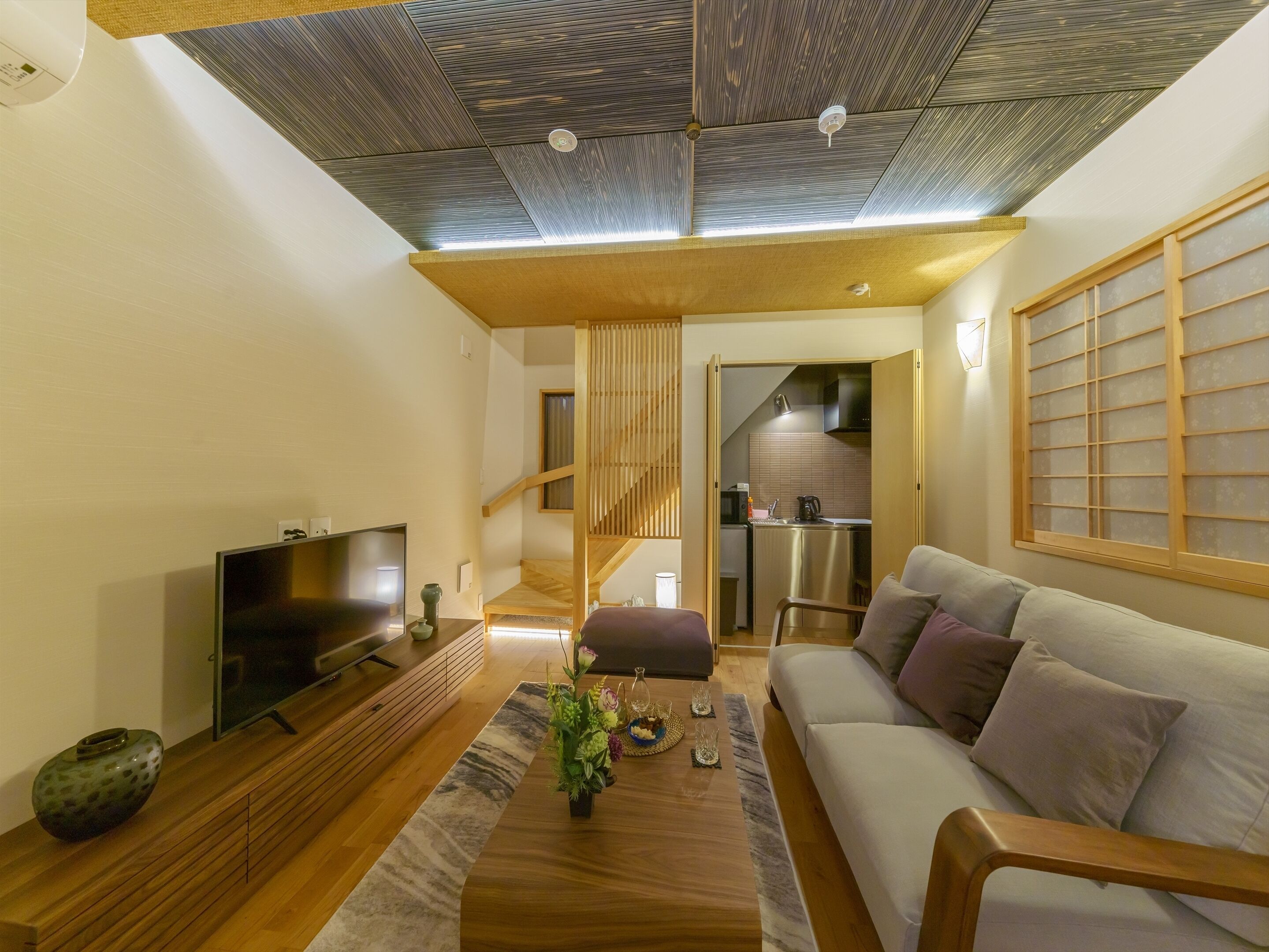 A modern living room with a flat-screen TV, wooden furniture, a sofa, and a kitchen in the background.