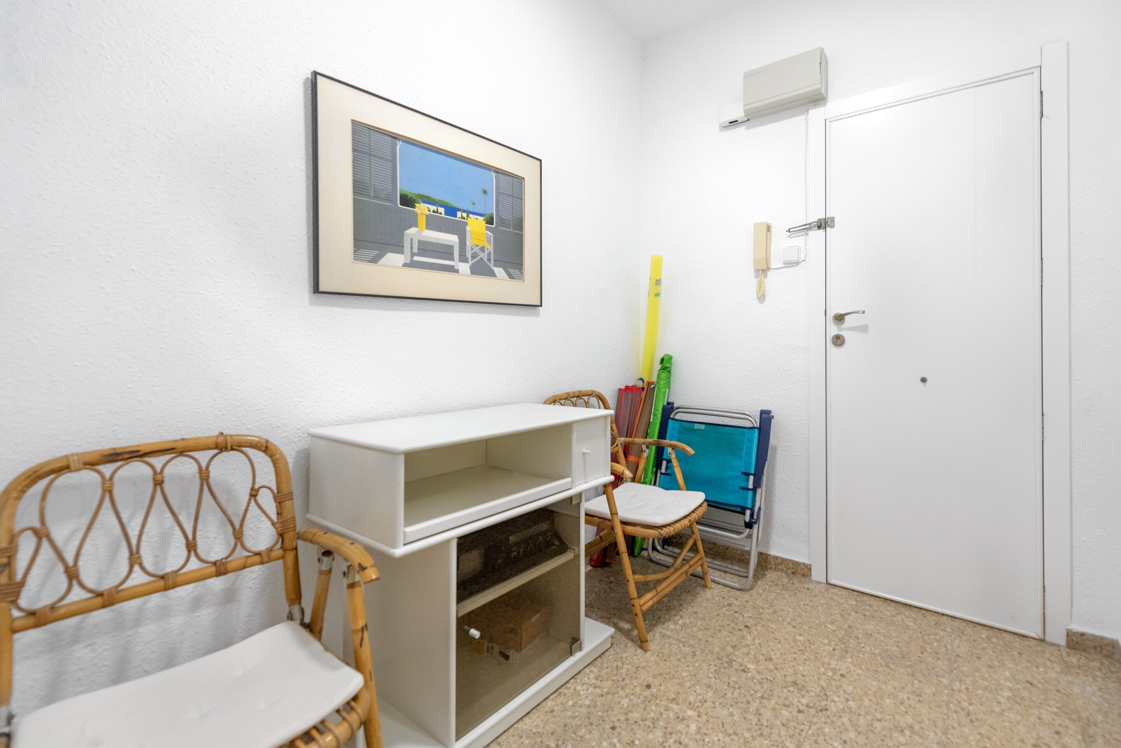 Una habitació amb una porta blanca, un quadre al mur, un armari blanc, una cadira de vimet i una cadira blava.