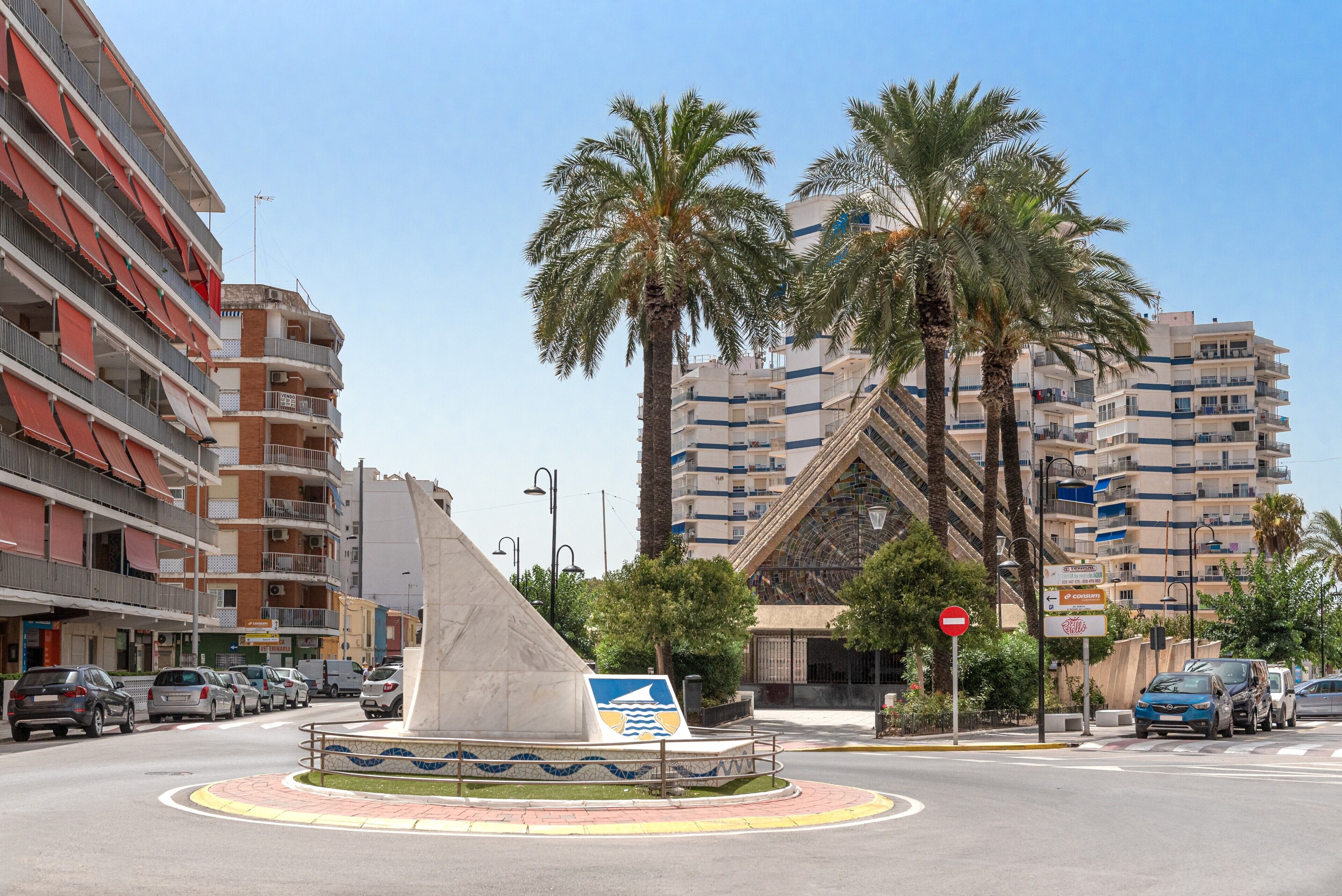 A multi-story building with balconies, a roundabout with a sculpture, palm trees, and a clear sky.