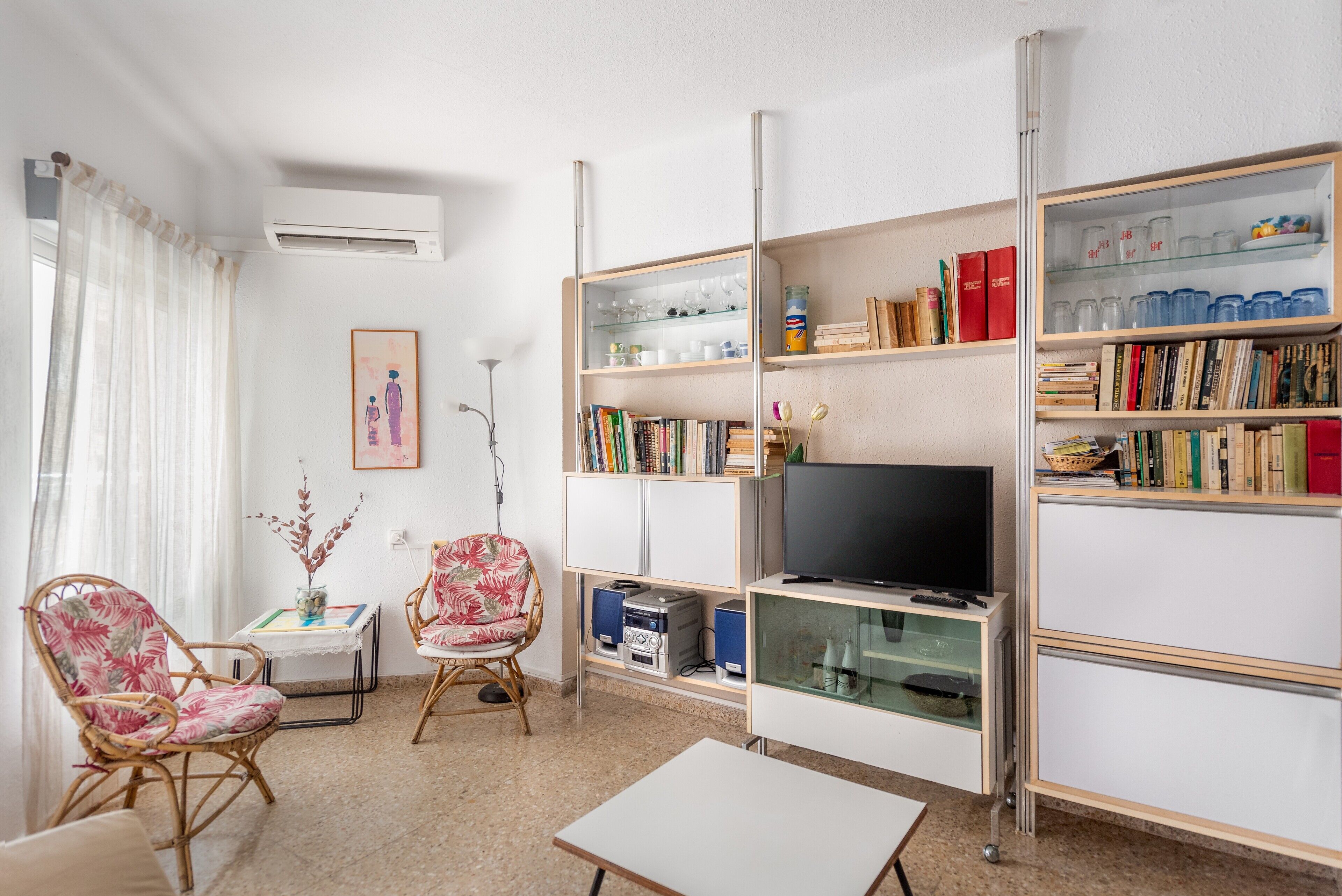 A living room with a TV, bookshelves, two wicker chairs, a small table, and an air conditioning unit.