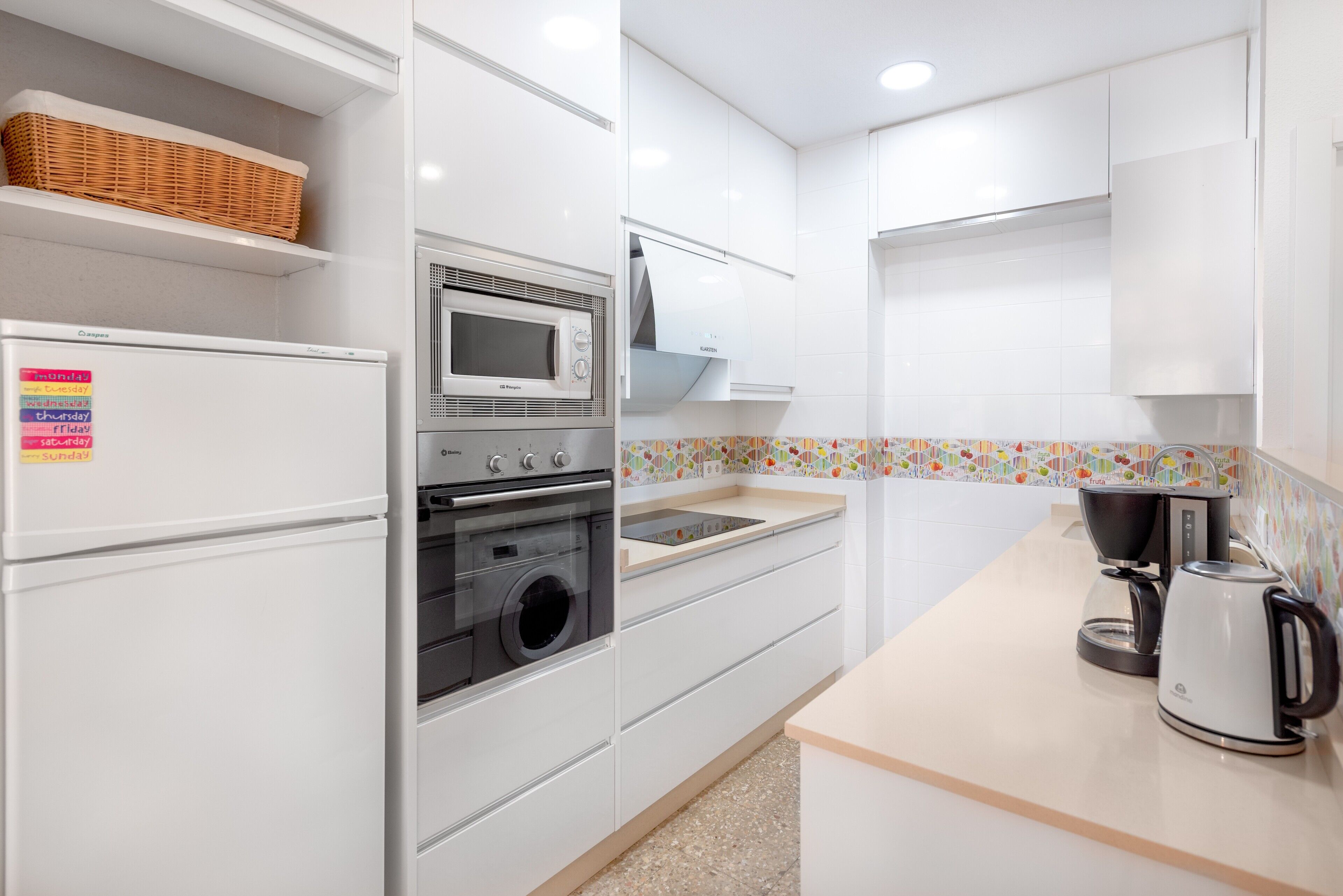 Cocina moderna con armarios blancos, microondas, horno, lavadora y cafetera.