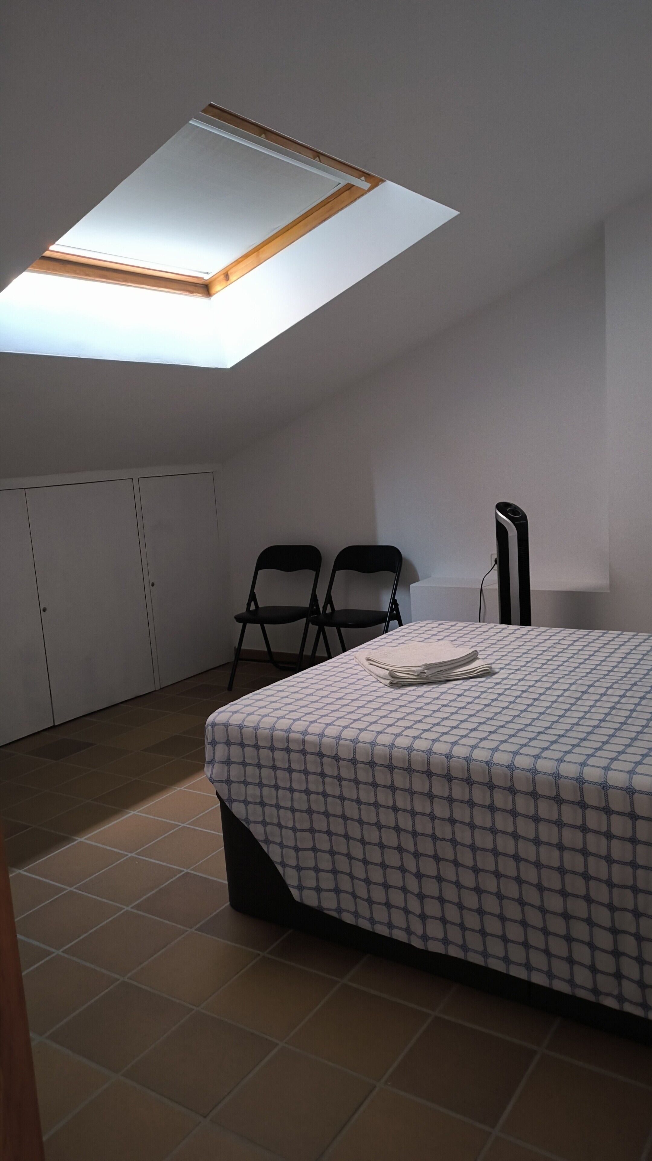 Una habitación con tragaluz, dos sillas, una cama con colcha estampada y un objeto alto y negro.