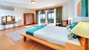A bedroom with a large bed, a wooden bench, a desk, a chair, a wardrobe, and a view through the windows. - Villa 'Albuar' with Sea View, Pool, Garden & Wi-Fi (São Rafael)