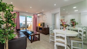 Una sala de estar moderna con sofá, mesa de comedor y sillas, televisor y un amplio ventanal con cortinas.