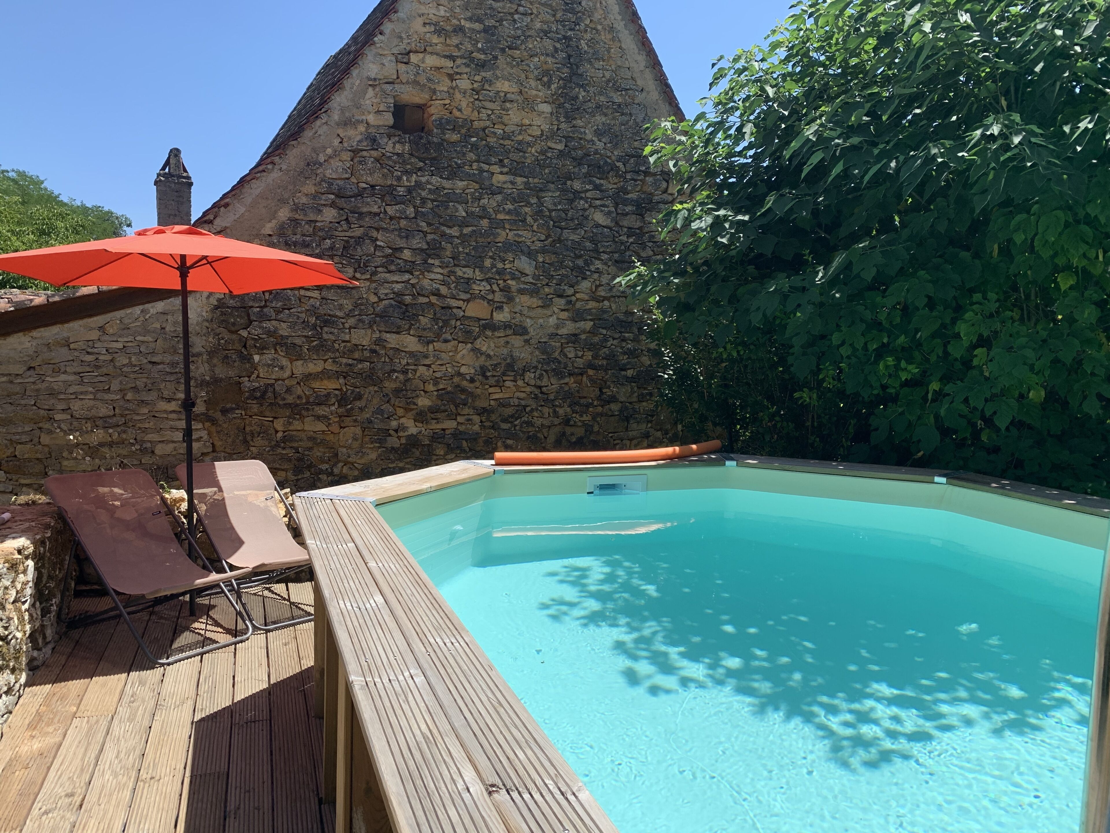 A swimming pool with a wooden deck and lounge chairs, surrounded by stone walls and greenery.