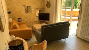 A cozy living room with a stone wall, a sofa, a chair, a TV, and a window with a view of greenery. - Authentic stone house in a typical village at the foot of a castle! (Berbiguières)