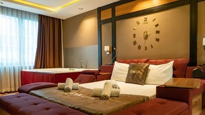 A hotel room with a large bed, a sofa bed, a wooden side table, a clock on the wall, and a window with curtains.