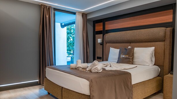 A modern hotel room with a large bed, a bedside table, and a view of greenery outside.