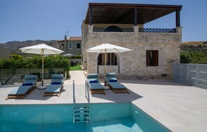 Ein Poolbereich mit Liegestühlen und Sonnenschirmen, ein Steinhaus mit Balkon und freie Sicht auf die umliegende Landschaft.