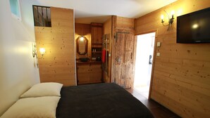 A bedroom with a bed, pillows, a wooden wardrobe, a mirror, a television, and a bathroom visible through an open door.