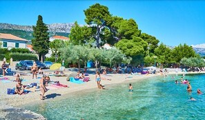 Una playa con agua cristalina, gente tomando el sol y nadando, y árboles al fondo.