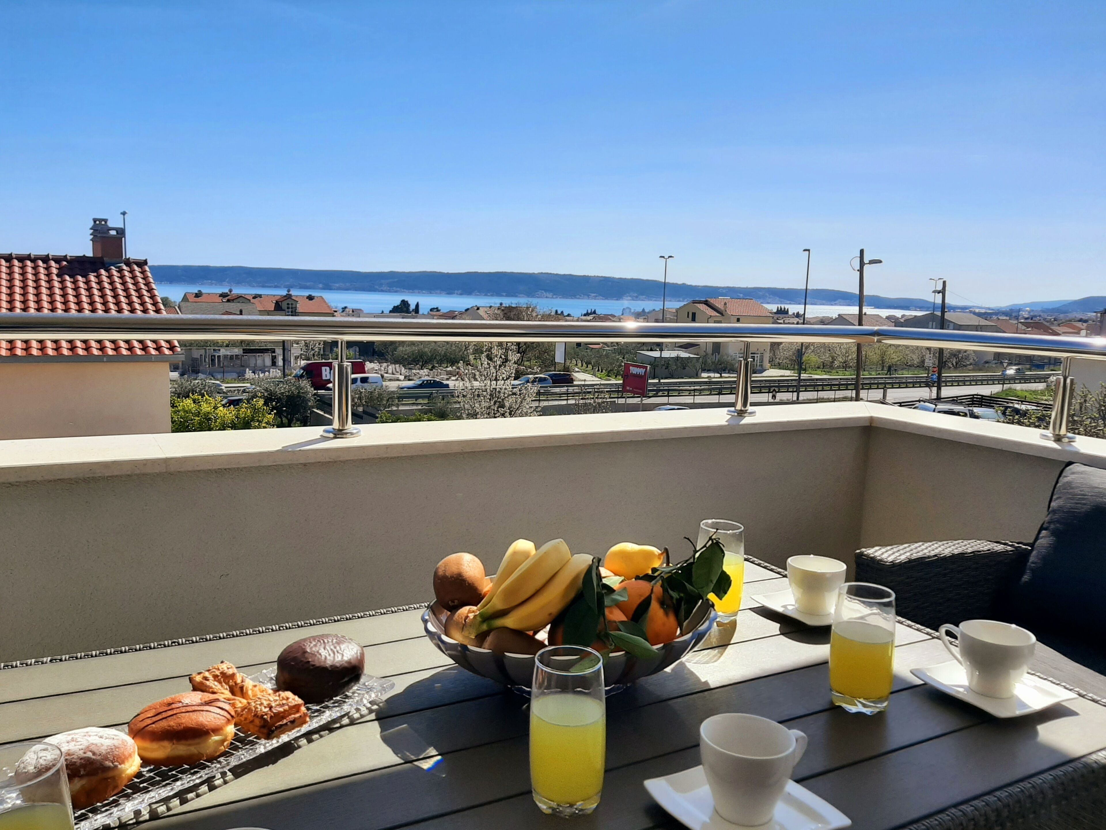 Un balcón con una mesa dispuesta para el desayuno, que incluye bollería, fruta y bebidas, con vistas panorámicas al mar y edificios.