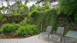 Un espace patio avec deux chaises en métal et une table, entouré d’une végétation luxuriante et d’un mur en pierre.