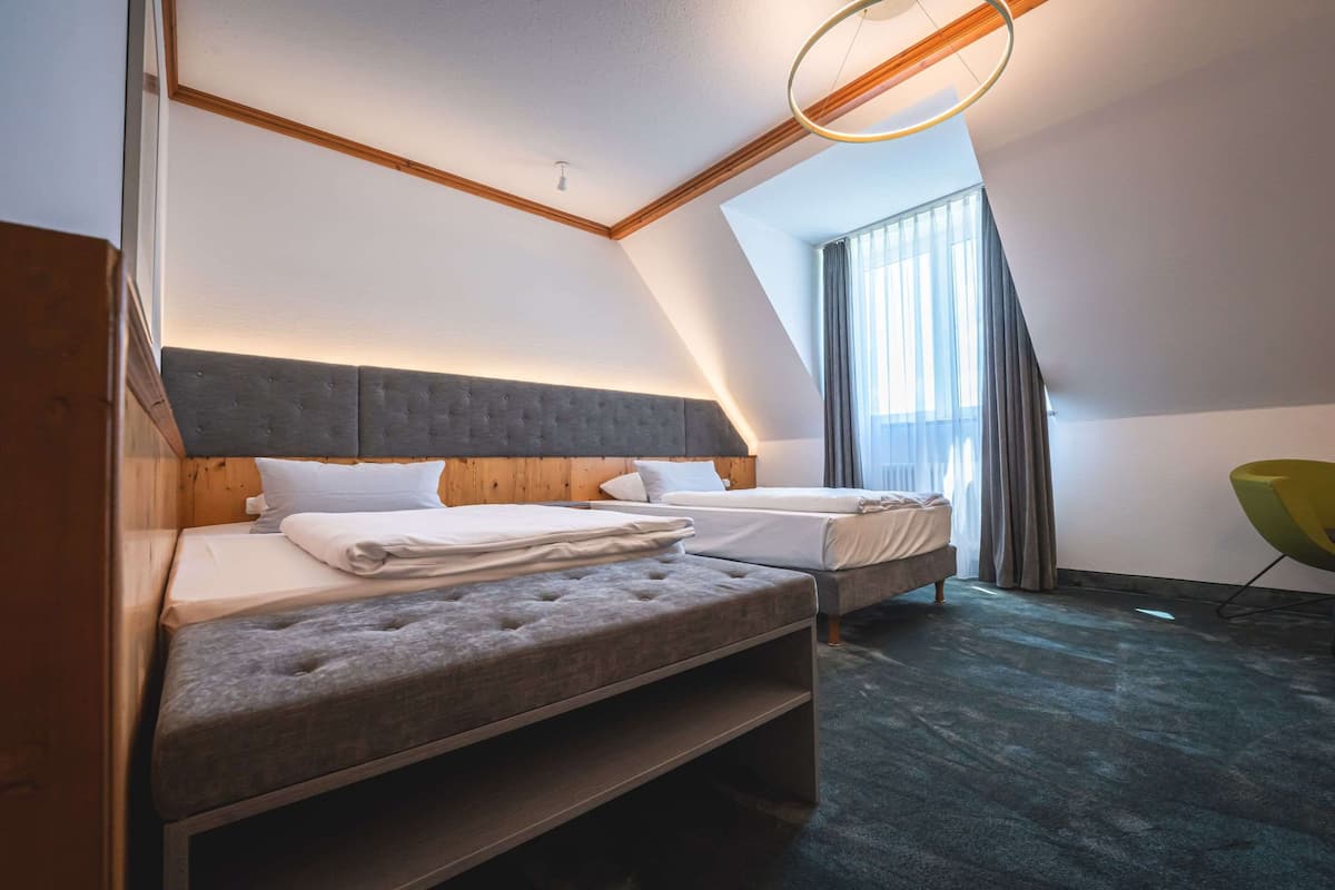 A modern hotel room with a sloped ceiling, two beds with grey upholstered headboards, a wooden accent wall, and a large window with curtains.