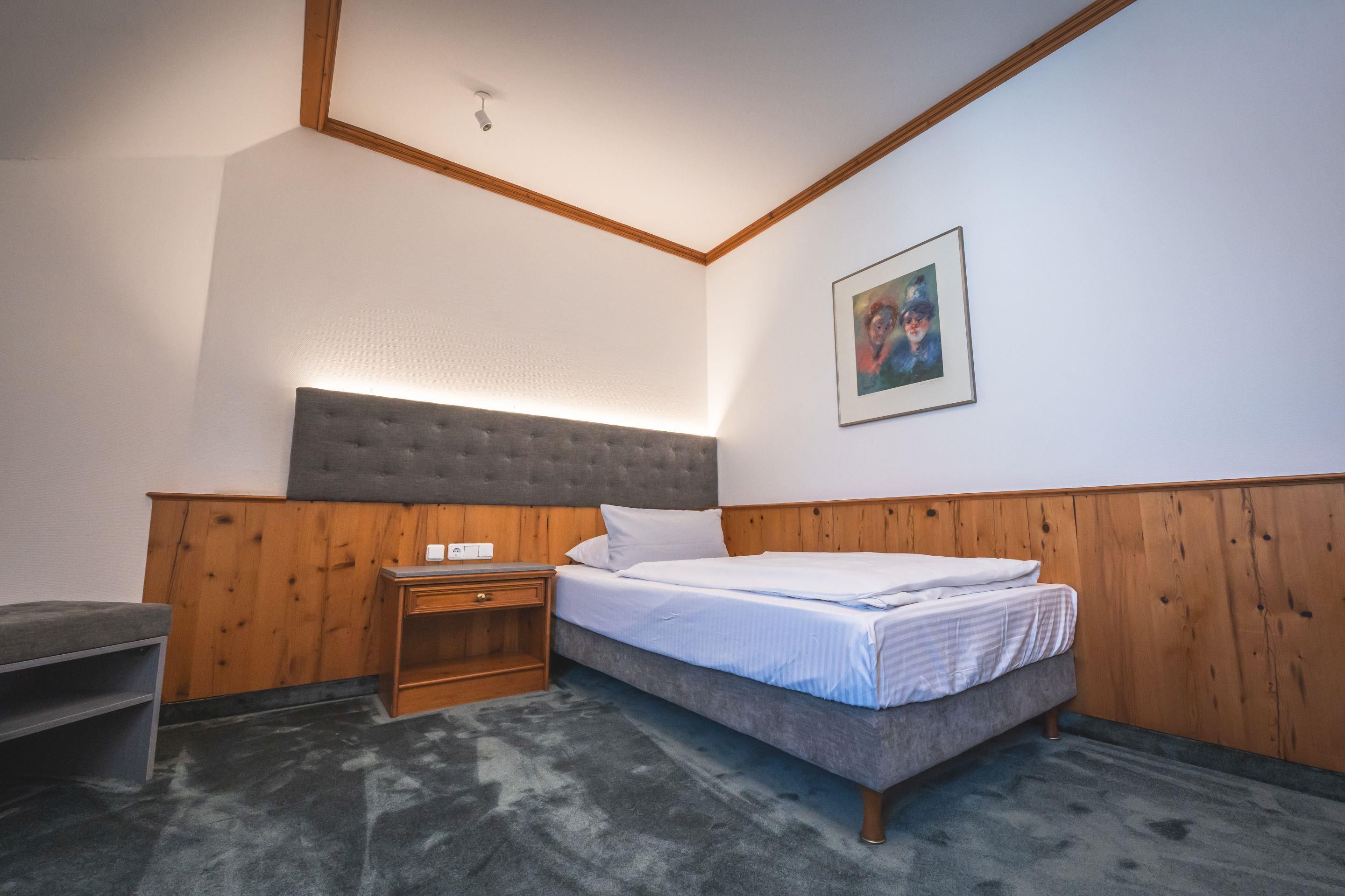 A bedroom with a bed, a nightstand, a wooden headboard, a wall-mounted picture, and a ceiling light.