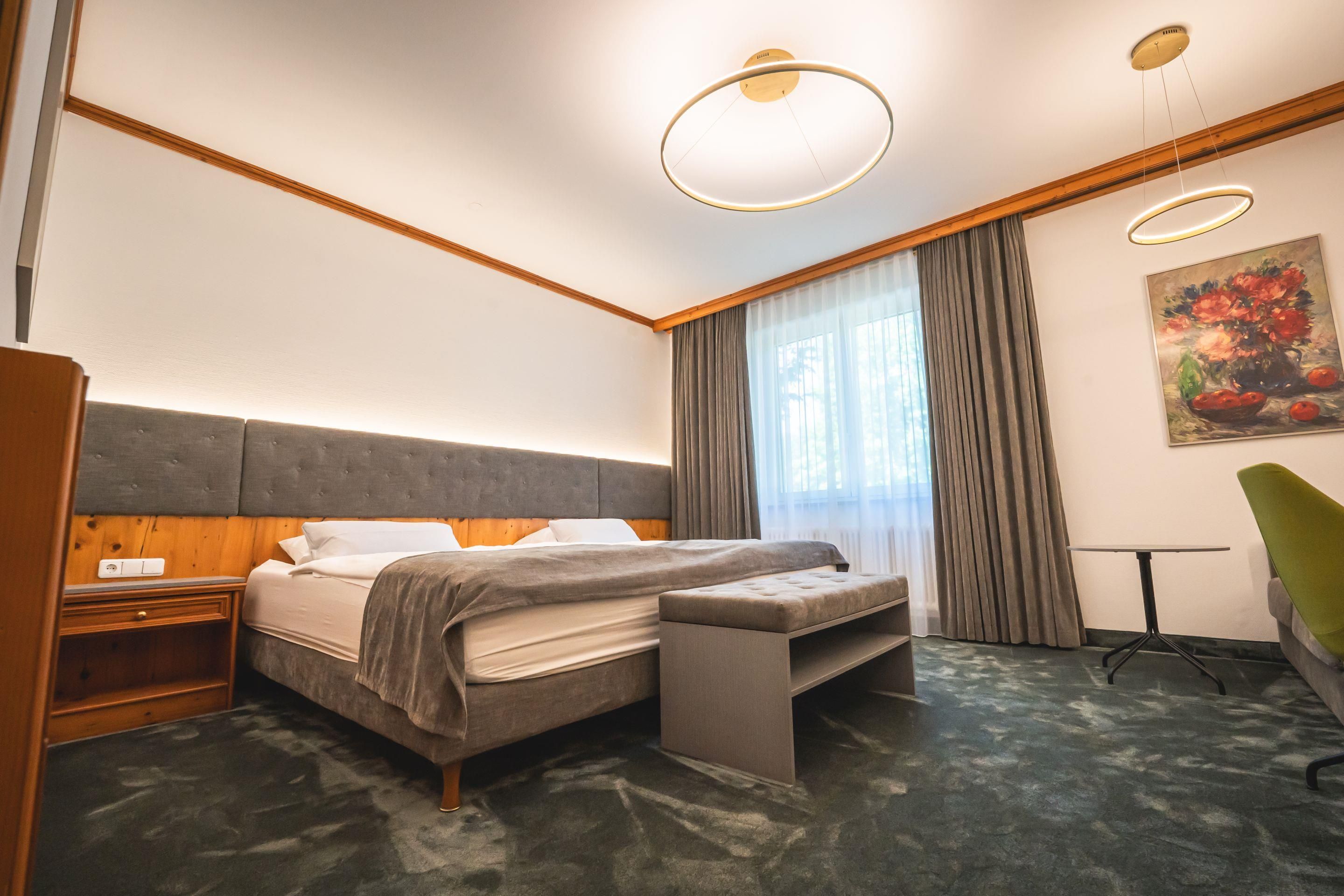 A hotel room with a bed, bedside table, a desk, a chair, a painting, and a window with curtains.