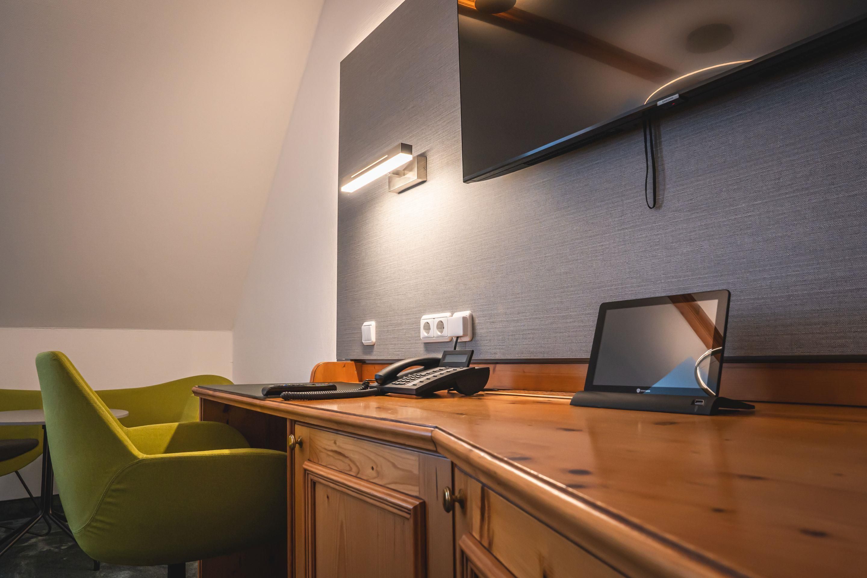 Een hotelkamer met een houten bureau, een groene stoel, een flatscreen televisie aan de muur en een laptop.