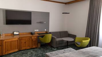 Ein Hotelzimmer mit einem wandmontierten Flachbildfernseher, einem Holztisch mit Telefon und Fernbedienung, einem grauen Sofa, zwei grünen Sesseln und einem grauen Hocker.