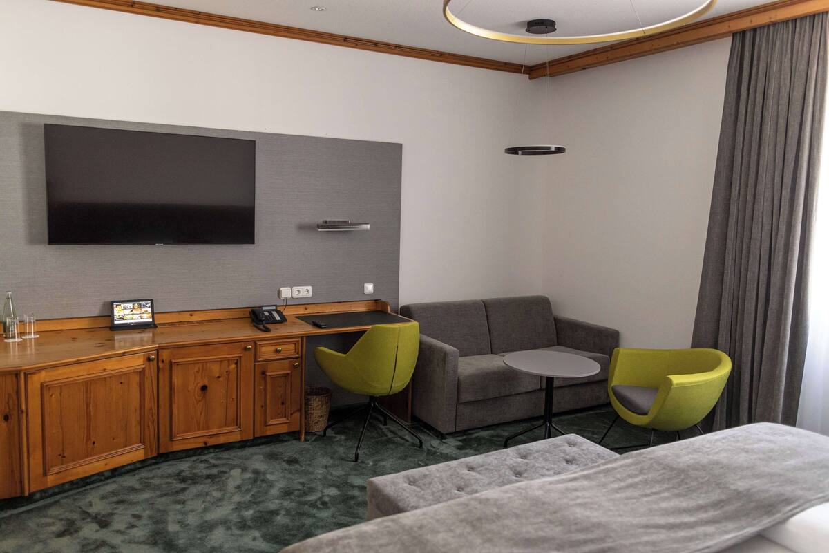 A hotel room with a flat-screen TV mounted on the wall, a wooden desk with a telephone, two green chairs, a grey sofa, and a small round table.
