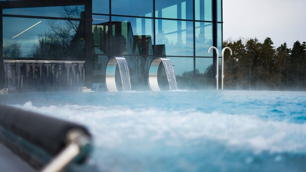 Un jacuzzi con vapor saliendo, una estructura metálica y un edificio moderno con grandes ventanales al fondo.