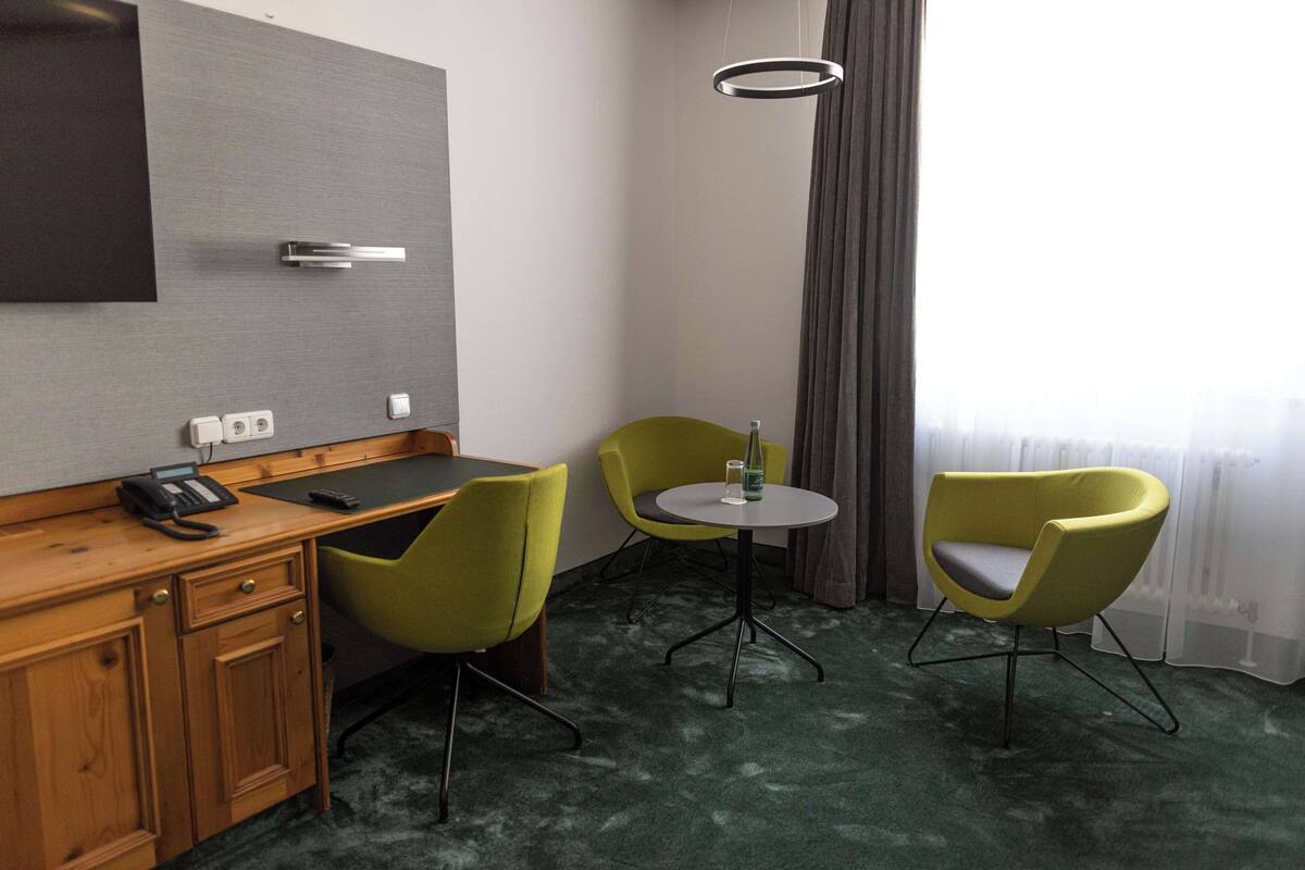 A hotel room with a wooden desk, two green chairs, a small round table, a telephone, and a wall-mounted TV.