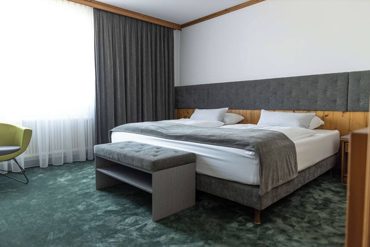 A modern hotel room with a large bed, a bench, a chair, and a view of the city through sheer curtains.