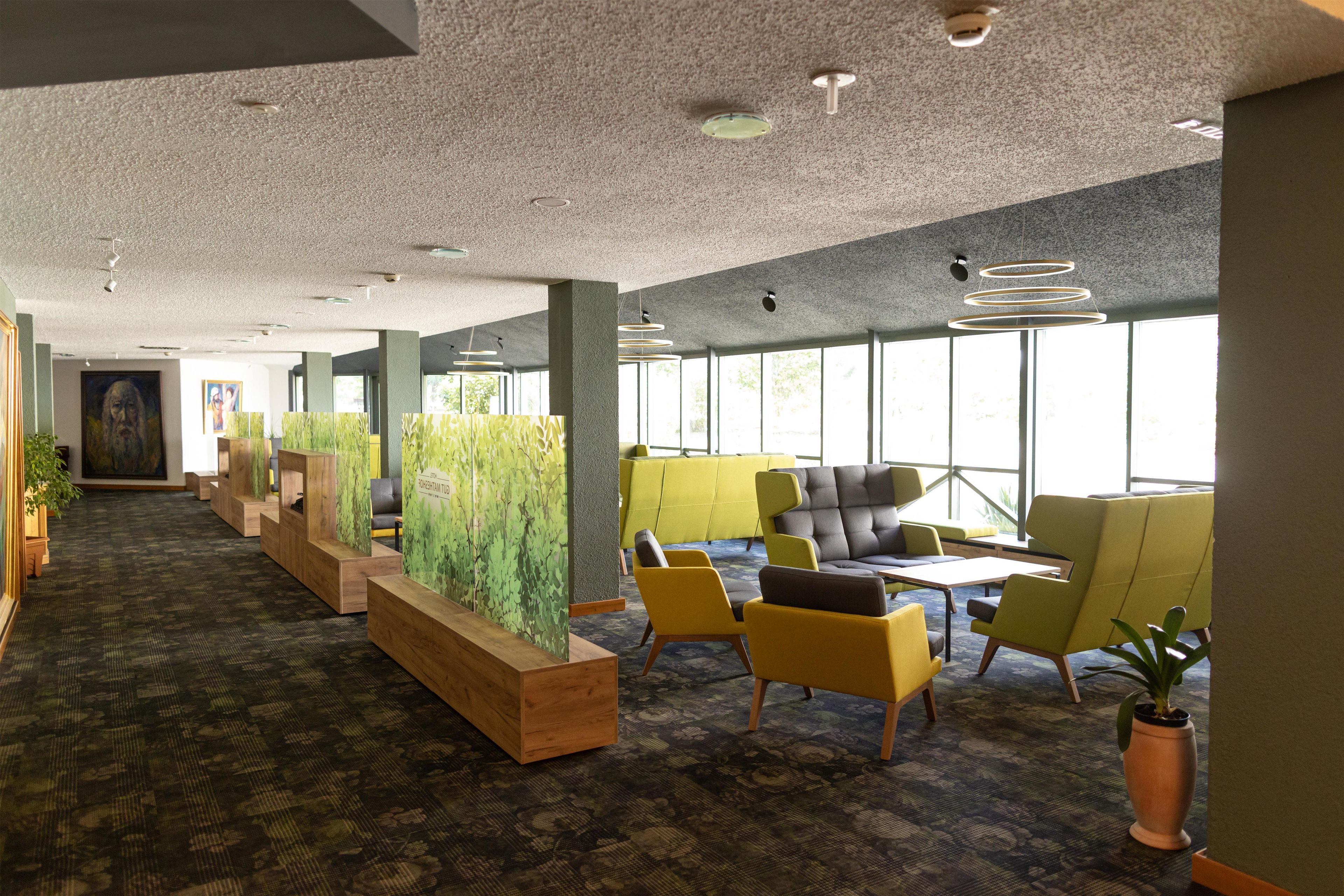 A modern office space with a long corridor, wooden planters, yellow and grey seating, and large windows.
