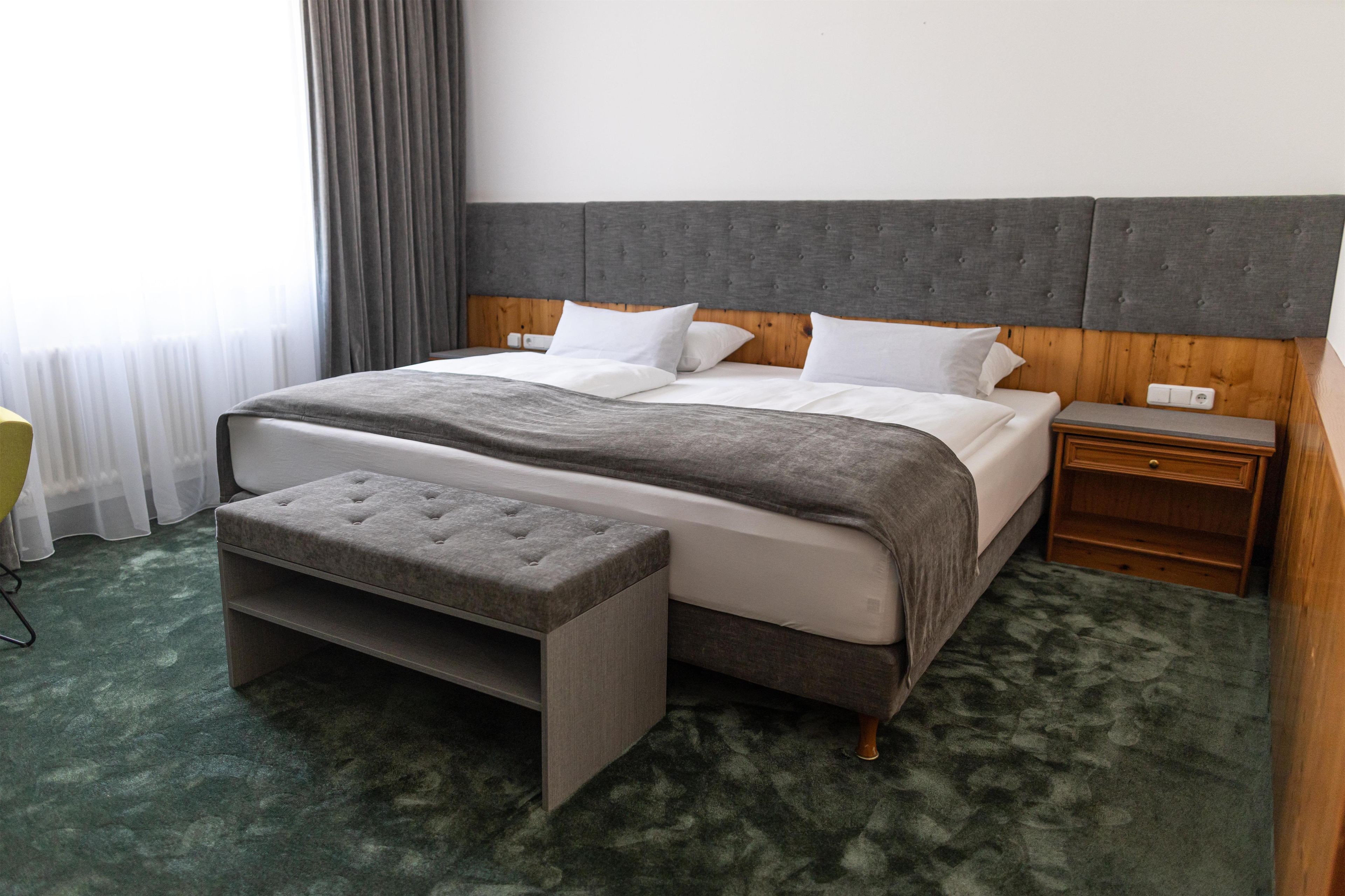 A hotel room with a bed, a wooden nightstand, a bench, and a window with curtains.