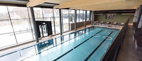 En stor inomhuspool med tydliga banor, omgiven av stora fönster som ger utsikt över omgivningen.