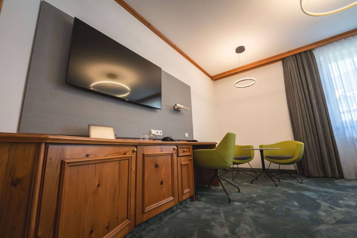 A hotel room with a wooden desk, two green chairs, a flat-screen TV mounted on the wall, and a large window with curtains.