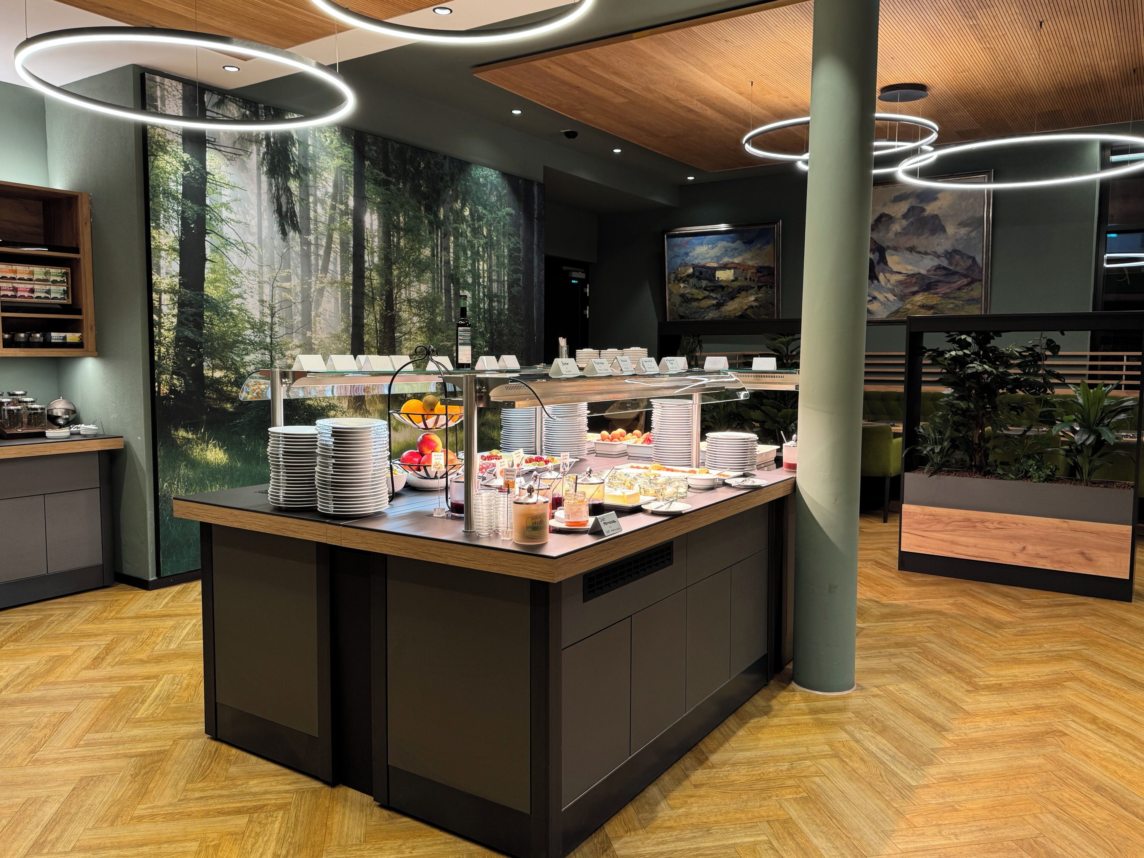 Een ontbijtbalk met diverse etenswaren, een koffiehoek en een groot muurdecoratie van een bos.