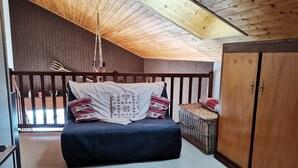 Un dormitorio con techo inclinado, una cama con manta blanca y almohadones estampados, un armario de madera y una cesta de mimbre.