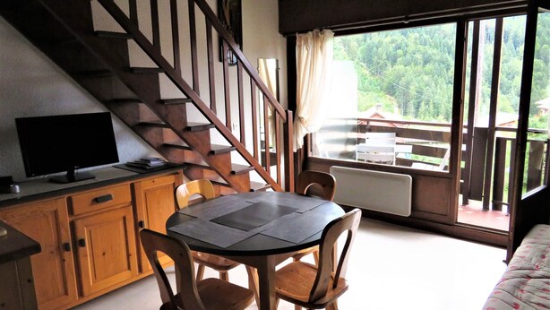 Un comedor con una mesa y sillas, un televisor sobre un mueble, una escalera y vistas a un bosque a través de un ventanal grande.