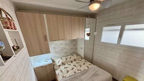 A compact room with a bed, a small desk, a shelf with decorative items, and a window.
