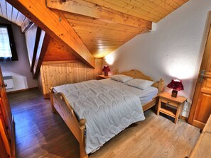 A cozy bedroom with a wooden ceiling, a bed with white bedding, a nightstand with a lamp, and a small wooden cabinet.