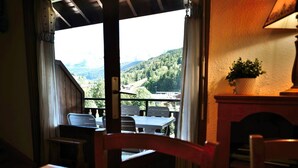 A dining area with a table and chairs, a view of a forested landscape through a window, and a lamp on a mantel. - Charming 2-Room Mezzanine Apt in Grand-Bornand Village, Sleeps 4, Balcony, Steps from Ski Lifts (Le Grand-Bornand)