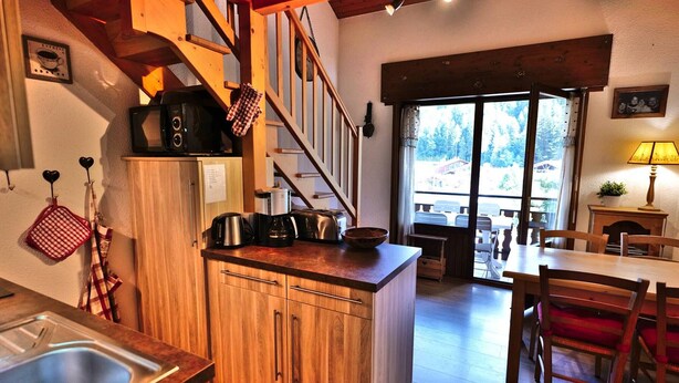 A kitchen with wooden cabinets, a coffee maker, and a microwave. There is a dining area with a table and chairs. - Charming 2-Room Mezzanine Apt in Grand-Bornand Village, Sleeps 4, Balcony, Steps from Ski Lifts (Le Grand-Bornand)