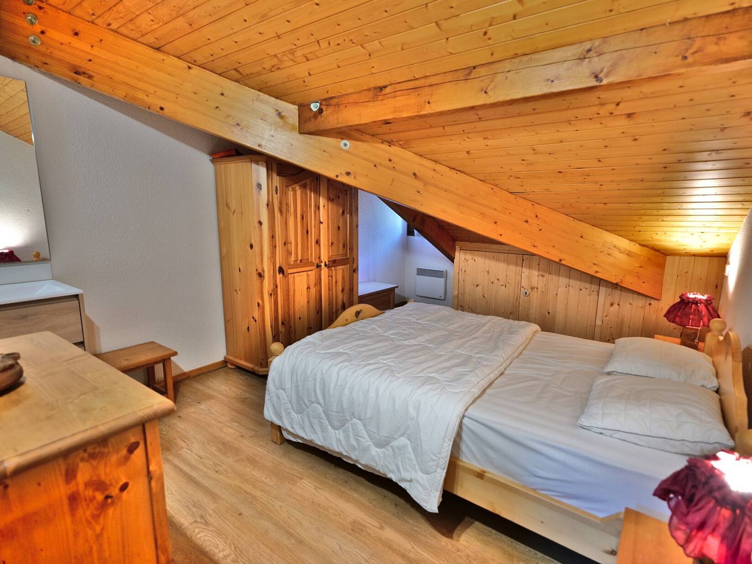 Ein Schlafzimmer mit schrägem Dach, Holzwänden, einem Bett mit weißer Bettwäsche, einer Holzkommode und einer roten Lampe.