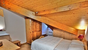 A bedroom with a sloped ceiling, wooden walls, a bed with white bedding, a wooden dresser, and a red lamp. - Charming 2-Room Mezzanine Apt in Grand-Bornand Village, Sleeps 4, Balcony, Steps from Ski Lifts (Le Grand-Bornand)