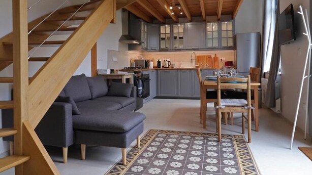 A modern interior with a staircase, grey sofa, wooden dining table, and patterned rug.