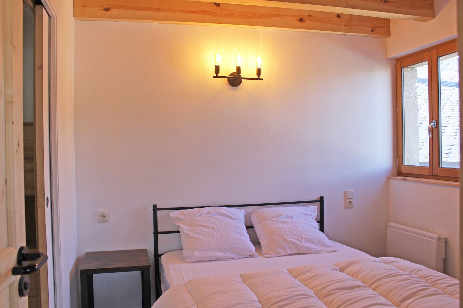 A bedroom with a bed, a bedside table, a wall-mounted light fixture, and a window.