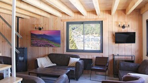 Un intérieur en bois comprenant un canapé, des fauteuils, une table, un tableau, un téléviseur et une fenêtre donnant sur un paysage.