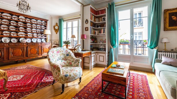 Un salon avec un fauteuil à motifs floraux, une table basse en bois, un tapis rouge, une bibliothèque et une vue sur l’extérieur grâce à de grandes fenêtres.