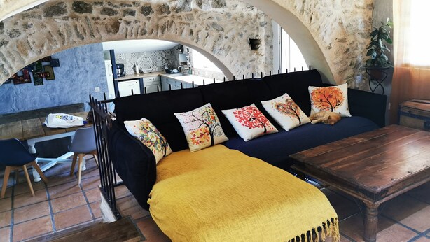 Ein gemütlicher Wohnbereich mit einem steinernen Bogen, einer Couch mit bunten Kissen, einem Holztisch und einer durch den Bogen sichtbaren Küche.